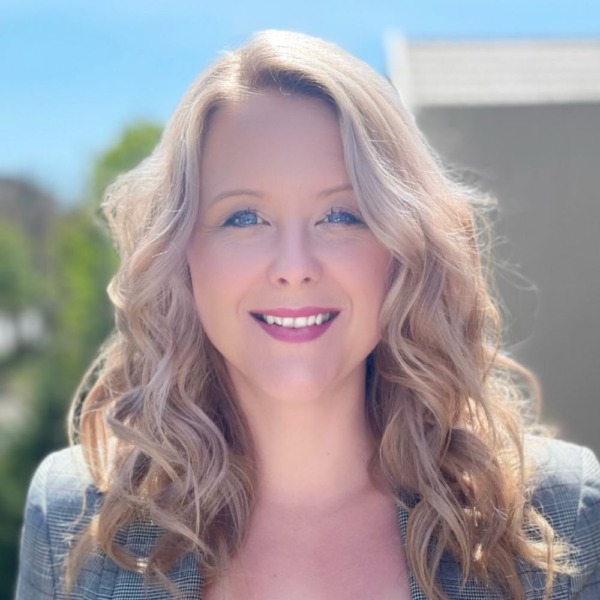 Woman with long curly blonde hair wearing a gray striped blazer and white top.