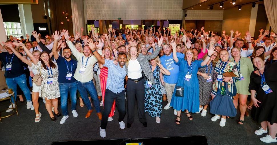 A group of event attendees standing in a conference hall smiling and waving at the camera