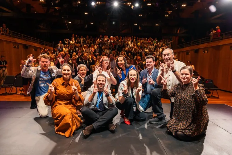 A group of speakers posing for a group photo in front of a large crowd in an auditorium.