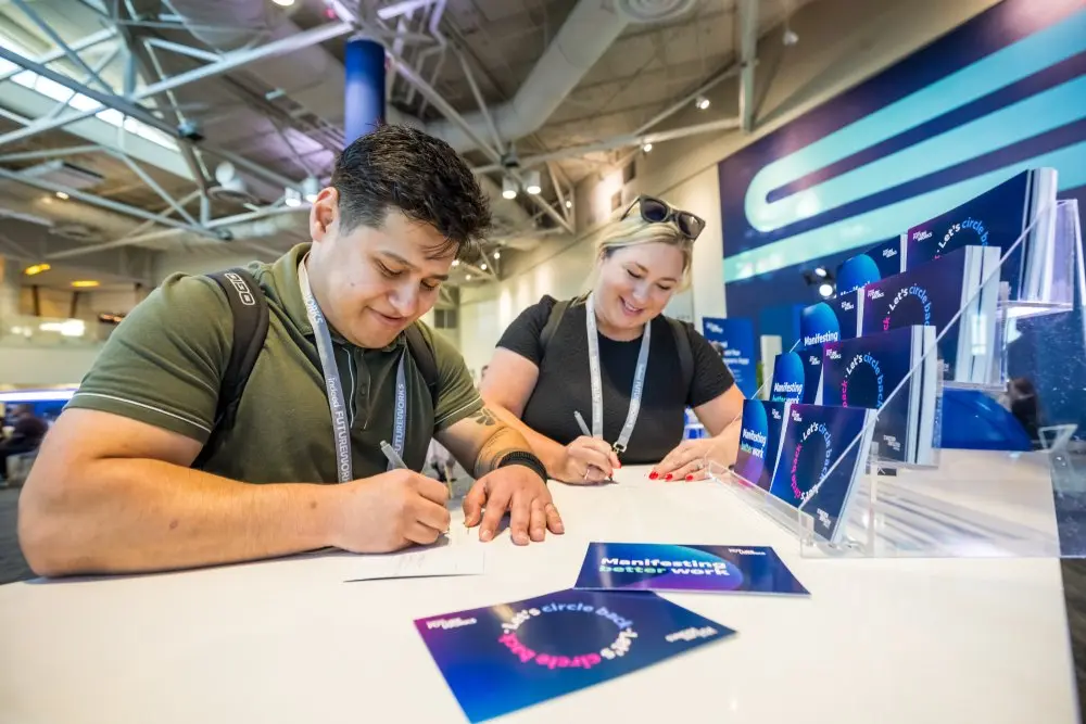 Two attendees at the Indeed FutureWorks event are writing on cards at a table. The man on the left, wearing a green shirt and rucksack, and the woman on the right, in a black shirt with sunglasses on her head, both look focused and cheerful. Displayed on the table are cards with phrases like 'Manifesting Better Work' and 'Let's circle back'. The background features modern event signage and lighting, adding to the professional atmosphere.