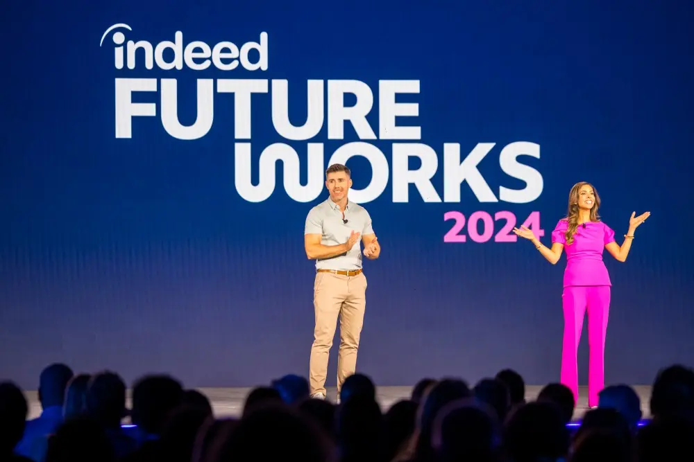 Two hosts stand on stage at the 2024 Indeed FutureWorks conference. The man on the left is dressed in a light shirt and khaki trousers, while the woman on the right is wearing a bright pink outfit. Both are engaging with the audience, with the event's logo and year displayed on a large screen behind them. Audience silhouettes are visible in the foreground.