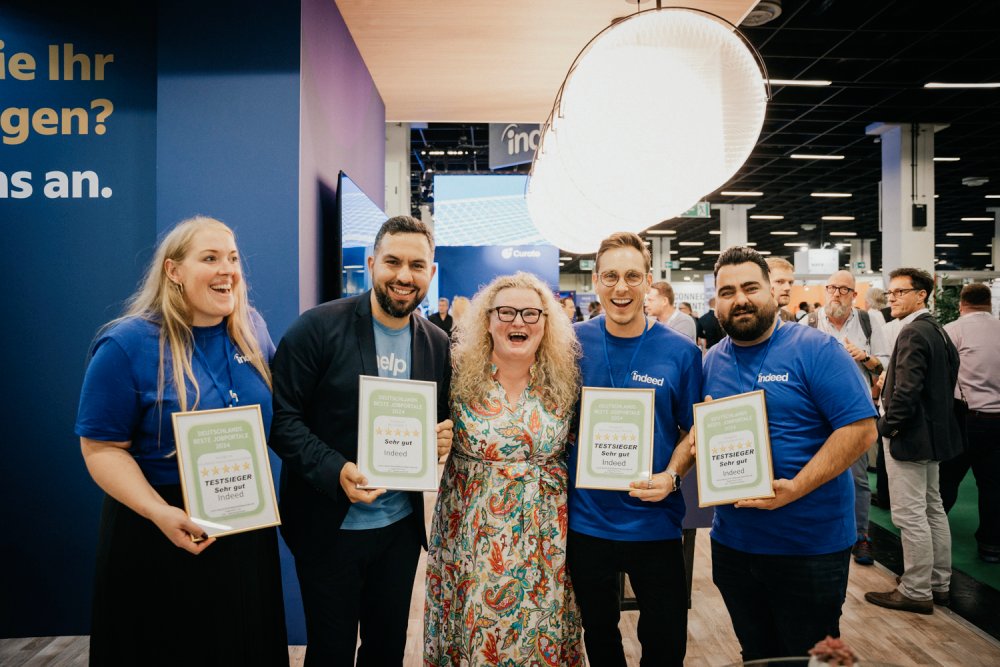 Five people, including four Indeed representatives in blue shirts, pose with certificates awarded to Indeed for being one of 'Germany's Best Job Portals 2024'. They are standing in a busy convention setting, smiling and celebrating the achievement.