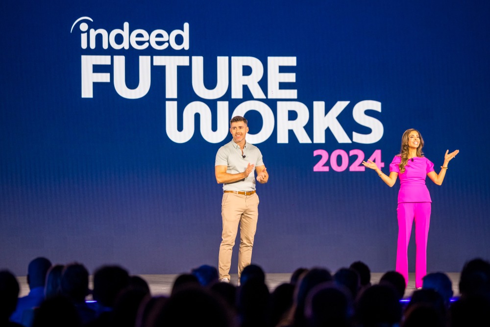 Deux hôtes sont sur scène lors de la conférence Indeed FutureWorks 2024, l'homme à gauche est vêtu d'un polo clair et d'un pantalon kaki, tandis que la femme à droite porte une tenue rose vif ; tous deux interagissent avec le public, avec le logo et l'année de l'événement affichés sur un grand écran derrière eux, les silhouettes du public sont visibles au premier plan