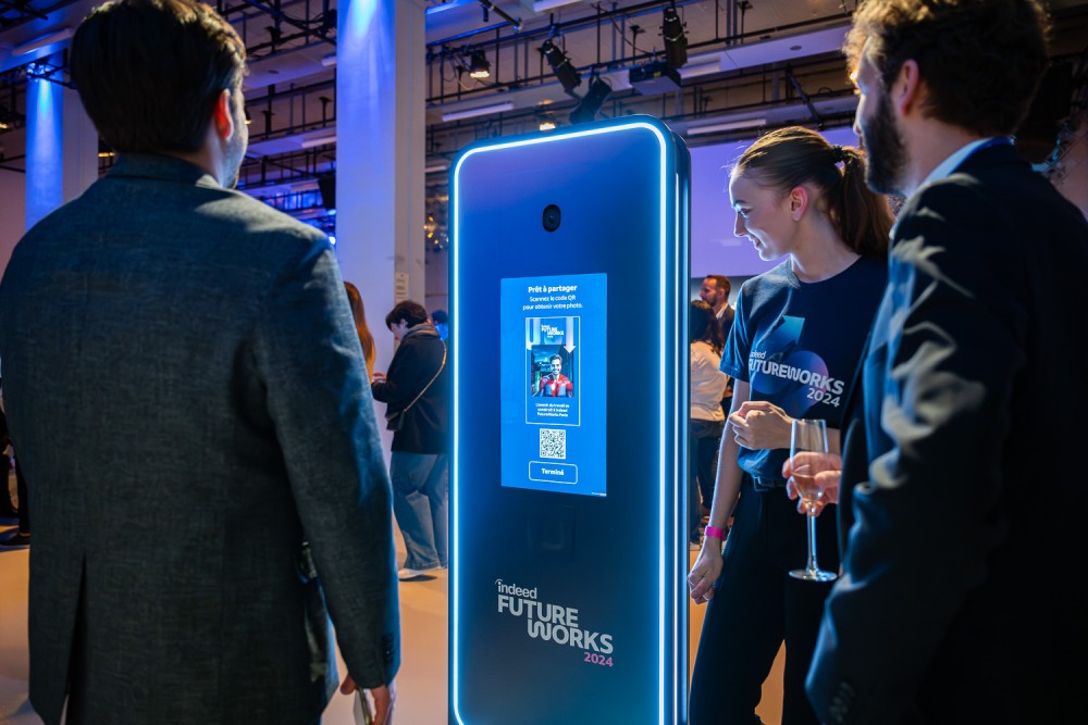 Trois personnes se réunissent autour d'un photomaton numérique futuriste aux bords bleu éclatant lors de l'événement Indeed FutureWorks 2024 ; une personne, vêtue d'un t-shirt de la marque, sourit en expliquant les caractéristiques du stand, l'écran affiche des instructions et un code QR pour partager des photos, à l'arrière-plan, les personnes participant à l'événement se trouvent dans un espace moderne et bien éclairé