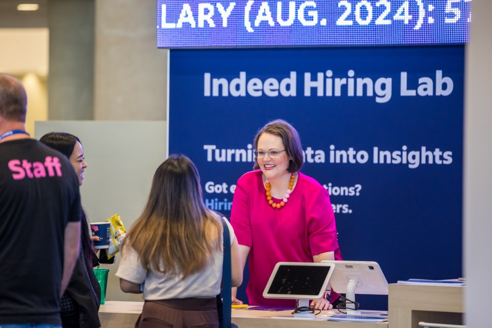 Une femme vêtue d'un haut rose sourit et discute avec des visiteurs sur le stand Indeed Hiring Lab ; un écran numérique au-dessus affiche « Indeed Hiring Lab » et des informations sur l'événement, tandis qu'un membre du personnel et d'autres participants écoutent à proximité