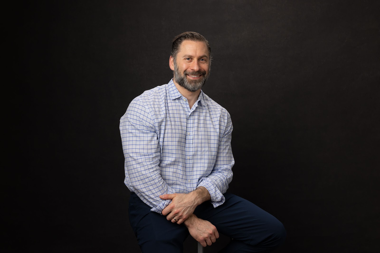 Tony Nelan's headshot. He is sitting on a stool, smiling. Wearing a light shirt and dark blue trousers.